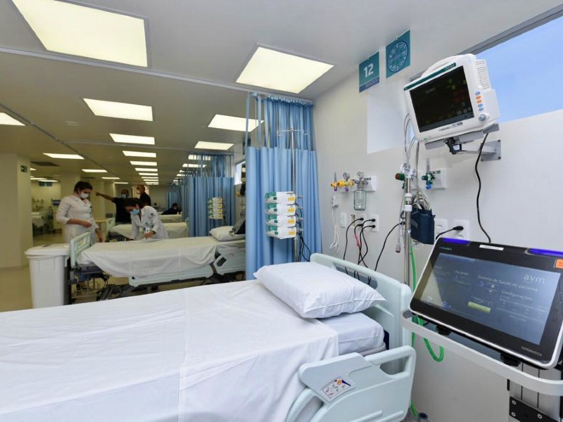 Foto dos novos leitos do Hospital Santa Catarina. Sala ampla com paredes beges à esquerda e azuis à direita. Em primeiro plano uma cama hospitalar com lençol e travesseiro brancos, dois monitores na cabeceira, ao fundo há mais quatro camas e seis pessoas uniformizadas trabalhando
